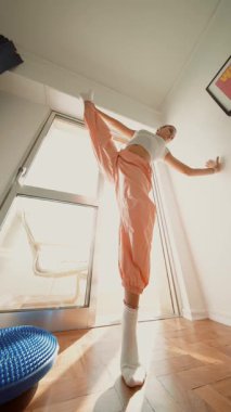 Beyaz çağdaş dansçı pencerenin kenarına döner, turuncu pantolon ve çoraplar hareket bulanıklığı yaratır, güneş ışığı yayılır.