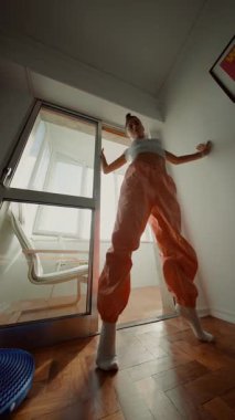 Beyaz model, güneşli kapı aralığında cesur poz veriyor, turuncu peruk pantolon ve sıradan çoraplar, uzun gölgeler bırakan doğal ışık.