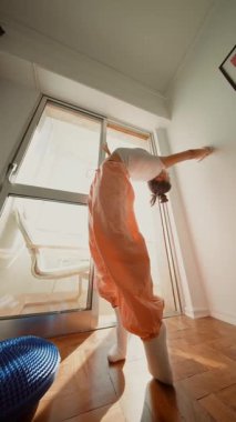 Beyaz sanatçı parlak pencerenin yanında arabesk tutuyor, turuncu pantolon ve çorapların içinde ince bir siluet, yumuşak sabah ışığı, odaklanmış