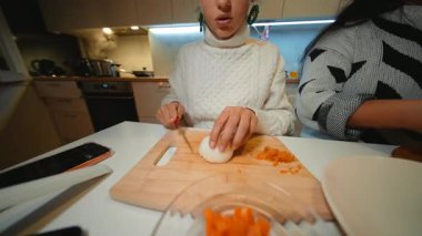 Kadın sıcak mutfakta soğan doğruyor, ev aşçısı beyaz süveter giyip soğan dilimliyor, yol arkadaşı da yardım ediyor, akıllı telefon.