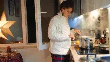 Beyaz kadın, gece kompakt mutfakta buharlı yahni tadıyor, kaşık dolusu baharatlı ve ayarlayıcı süveter deniyor.