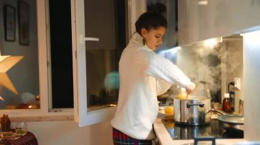 Beyaz kadın gecenin bir yarısı sade mutfakta haşlanmış tencereyi karıştırıyor tencerede kaynamış çorbayı kontrol ediyor, kablo örgü kazak giyiyor.