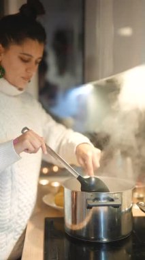 Beyaz kadın sıcak çorbayı tencereden kaseye atıyor, dikkatli hareketler, et suyu ve bitkiler görünür, rahat mutfak ışıkları.