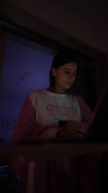 Laptop vitrininde genç bir kadın loş neon parıltılı telefonu kaydıran, rahat süveter giyen, yumuşak pembe ile akşam çalışması yapan.