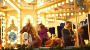 Akşam atlıkarınca ışıkları gösterişli atlar ve biniciler, çelenkler ve ön planda şenlik kurdeleleri ile hareketli bir panorama yaratır.,