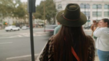 Şapkalı bir kadın kavşakta bekliyor. Sırt çantalı bir yayanın arka siluetini gösteriyor.