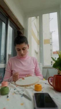 Genç bir kadın parlak pencerede kumaş dikiyor, nakış işlemeli, renkli iplik, masada akıllı telefon, konsantre eller.,
