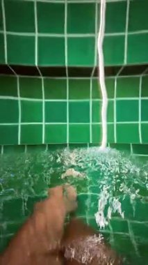 Bacakları yeşil fayanslı küvete gömülü, musluktan gelen yumuşak akış ayak etrafında kabarcıklar yaratır, yakın çekim dalgalanmaları yakalar.,