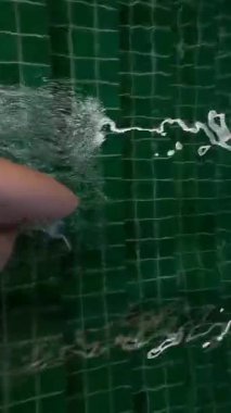 Beyaz kadın yeşil havuzda su sıçratıyor, ayakları tekmeliyor, selfie kolu görünüyor, enerjik dalgalar, spa ortamı, samimi eğlence.