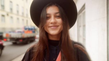 Şehir caddesinde gülümseyen bir kadın, siyah şapkalı ve uzun esmer saçlı yakın plan portresi, sıcak bokeh arka plan, samimi kafa eğik,