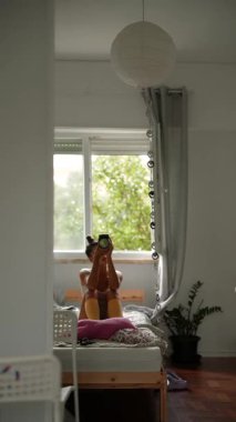 Yatakta gün ışığı altında selfie çeken kişi, gri perdeler ve kağıt fener, yatağın yanında saksı bitkisi, pembe