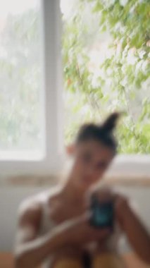 Pencerenin yanında kendi portresini yapan kadın, yumuşak yeşil arka plan, oturma pozisyonu, kamera göğse yakın, nazik gülümseme, sıcak yansıtıcı.