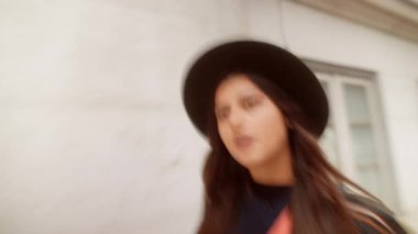 Düşünceli kadın şehir caddesinde yürüyor, şapka ve çapraz ceset torbası, sessiz tonlar, park etmiş arabalarla dar kaldırım, yansıtıcı.