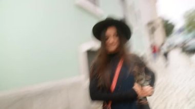 Şapkalı bir kadın Pastel caddesinde yürüyor, telefonu kontrol ediyor, kaldırımda kaldırım kenarında yürüyor, önünden geçen dükkanlar bulanık.,