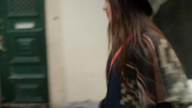 Şapka ve desenli montla dükkanların önünden geçen bir kadın, elinde telefon, hayat dolu sokak geçmişi, kendine güvenen bir ifade, dinamik