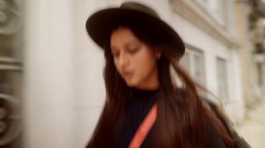 Bulanık kadın şehir caddesinde aceleyle yürüyor, hızlı el hareketi, şapka ve çapraz ceset torbası çerçevede, park halindeki enerjik aceleyle geçiyor.