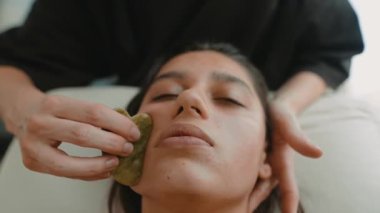 Asyalı bir kadın yeşim taşıyla dudak bölgesi heykeli yapıyor. Estetisyen, ağız ve çene boyunca yukarı doğru ölçülmüş darbeler uyguluyor.