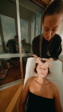 Yüz ritüeli kapanıyor, pratisyen, alnı ve çenesi boyunca hafif vuruşlarla bitiriyor, balkon güneş ışığı, gevşemiş istemci,