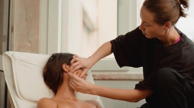 Beyaz kadın yüz lenfatik drenajı alıyor, terapist çene ve kulak boyunca hafif darbeler kullanıyor, mindere yaslanıyor, yavaş