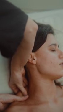 Kafkas kadın boyun masajı yaptırıyor, terapist trapezius üzerinde sabit parmaklar kullanıyor, beyaz yatağa yaslanıyor, yumuşak doğal ışık,
