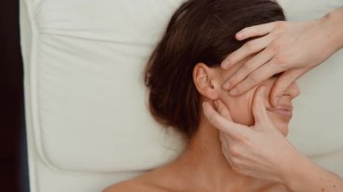 Beyaz kadın miyofasiyal çene boşaltımı geçiriyor. Pratisyen yüz fasyasını ölçülü basınçla geriyor.