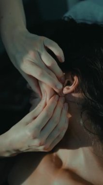 Yakın plan beyaz kadın, güneş ışığı altındaki yatak odasında kulak aküpresi yaptırıyor, eller kulak kenarına ve boynuna hafif basınç uyguluyor, sakinleştiriyor.