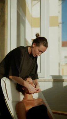 Sunlit stüdyo estetisyeni yüz bakımı yapıyor. Arkasına yaslanan müşteri nazikçe el masajı yaptırıyor.