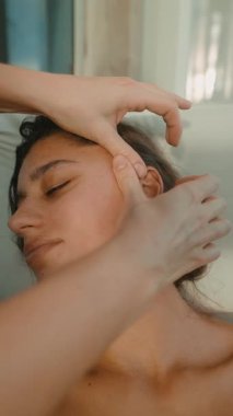 Yatağa yakın beyaz kadın nazik avuç içleri ve parmak uçlarıyla iyileştirici kulak masajı yapıyor. Yumuşak perde arkası ve gevşemiş.