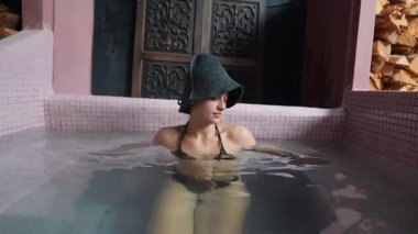 Jakuzide aşağı bakan bir kadın, keçeli bir şapka çerçevesi, sessiz bir ışık ve sakin bir su, içe dönük bir portreyi ve sakin bir spayı vurguluyor.