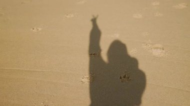 Gölge silueti kumlu sahilde, el kaldıran kişi barış işareti, dağınık ayak izleri, altın güneş ışığı, sakin