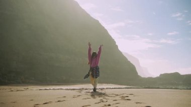 Sahilde büyük pembe kazakla dans eden Asyalı bir kadın, kolları havada ve falsolu, beline ekose gömlek bağlanmış.,