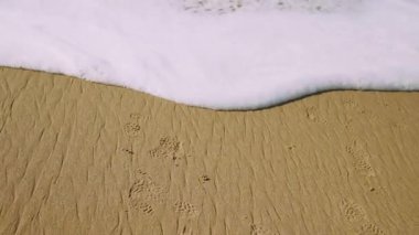 Köpüklü sahil şeridinde ıslak kumda ayak izleri, görünür ayak parmağı, kabarcıklı köpük geri çekiliyor, desenli kum detayı, yumuşak gel-git hareketi