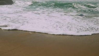 Kumlu sahilde okyanus dalgaları, yumuşak köpüklü kıyılar, yeşil mavi dalgalar, yavaş ritmik dalgalar, bulutlu ışık, sakin bir hava yaratıyor.