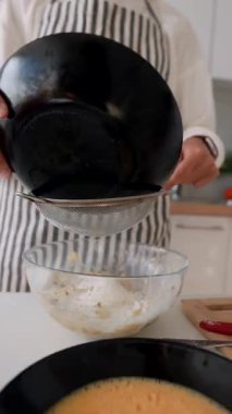 Ev mutfağı pişirme sahnesi, cam kaseye un eleme, çizgili önlük, düzgün eller, ince elek, dökme demir un