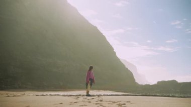 Kadın güvenli bir şekilde kumların üzerinde yürüyor, kameraya doğru düz bir adım, parlak güneş ışığı ve geniş bir uçurum arkaplanı, gülümseme ve neşeli bir kol.