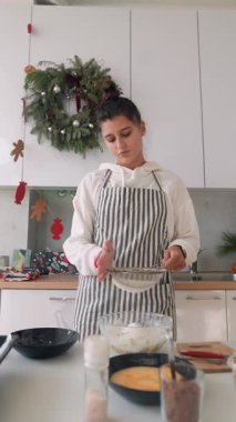 Mutfak kadını cam kaseye un karıştırıyor, tezgah üzerinde elek, yumurta kasesi ve baharatlar ön planda, çelenk tatili dekoru.,