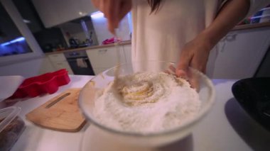 Cam kasede karıştırıcı hamur, unu içine katlayan kaşık, tezgahta sabitleyici kase, sıcak mutfak ışığı.