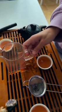 Küçük bardakta kehribar çayı yudumlayan ve koklayan bir kadın tepsiyle, süzgeciyle ve çaydanlığıyla yakından tatma anı, duyusal odaklanma.