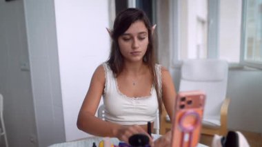 Pencere kenarına rimel süren yaşlı kadın, akıllı telefon aynası ve kompakt, sakin odaklı güzellik rutini ile güneşli gösteriş sahnesi,