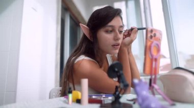 Pencerede hevesle makyaj aletleri gösteren ve rimel testi yapan genç bir kadın, görünür elf kulakları, telefon ve mikrofon yakalama