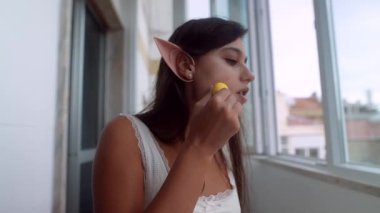 Pencerede duran genç bir kadın süngerle fondöten sürüyor, elf kulakları görünür, telefon tripoda ve mikrofon kaydına bağlı, yumuşak doğal.