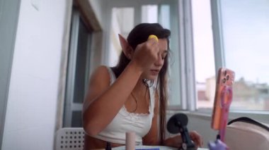 Pencerede saç ayarlayan ve iplikleri güvene alan genç kadın, elf kulakları detayı, telefon ve mikrofon çerçevede, sakin odaklı hazırlık