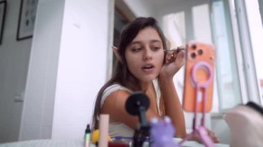 Balkonda genç bir kadın. Göz farını fırçayla harmanlıyor. Kulak aksanı, telefon ve mikrofon ön planda.