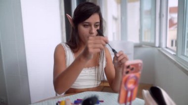 Pencerede genç bir kadın sabit elle göz kalemi sürüyor, elf kulakları detayı, telefon teçhizatı ve mikrofon yakalama gösterisi, anlamlı.