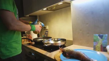 Kapalı soba tavası aşçısı yağ ve baharat ekler, eldivenli eller paslanmaz tavaya yağ döker, bitişik tavada indüksiyon hob 'u ısıtır.,