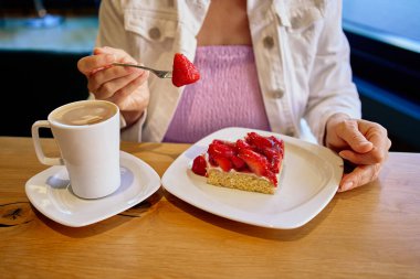 Sıcak bir kafede çilekli kek ve kahveden hoşlanan bir kadın. Taze çilekli çatal, beyaz tabakta tatlı ve sıcak kahve fincanı tutan ellerin yakın çekimi. Yaşam tarzı anı, tatlı mola, rahatlamış kafe atmosferi.