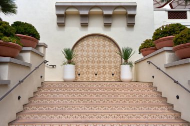 Şablonlu seramik merdiven, kemerli duvar oyuğu ve büyük saksı bitkileriyle zarif Akdeniz tarzı giriş. Yumuşak gün ışığında simetrik mimari sahne, rafine tasarım, lüks yaşam tarzı ve Güney Avrupa estetiği sergileniyor.