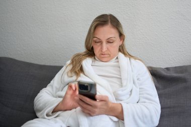 Orta yaşlı bir kadın, evinde kanepede dinleniyor, beyaz bornoz giyiyor ve akıllı telefon kullanıyor. Günlük teknoloji kullanımını, boş zamanı ve sessiz odaklanmayı gösteren rahat bir ev hayatı sahnesi..