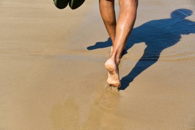 Sahildeki ıslak kumsalda yalınayak yürüyen biri. Bacakları ve ayakları doğal ışık ve gölgeyle yakın çekim. Yaz tatili, özgürlük, rahatlama, seyahat, deniz kenarı yaşam tarzı ve farkındalık kavramı..