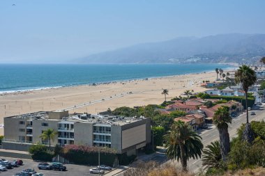 Okyanus dalgaları, palmiye ağaçları, konut binaları ve kıyı yolu olan geniş kumlu bir sahil manzarası. Güneşli bir gün, uzak dağların olduğu manzaralı bir kıyı şeridi boyunca. Deniz kenarında yaşama, seyahat yeri, sahil hayatı kavramı
